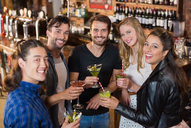 Group of friends standing together at a bar, holding various cocktails and smiling at the camera. Ideal for use in advertisements for nightlife, social events, bars, and restaurants. Perfect for illustrating themes of friendship, celebration, and leisure activities.