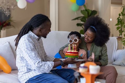 African american family celebrating child birthday on sofa with rainbow cake and balloons