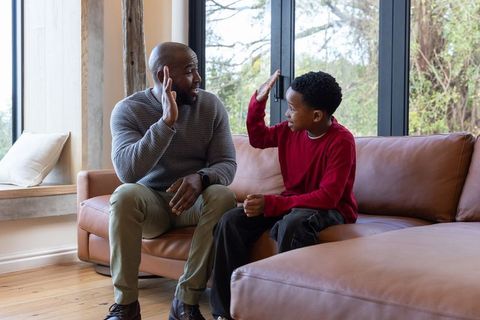 African american father and son sharing high five on tan leather sofa in bright living room