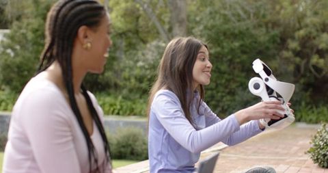 Female friends testing white vr headset outdoors on campus courtyard, sharing tech moment