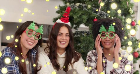 Three friends wearing christmas tree glasses and festive hats, laughing by decorated tree