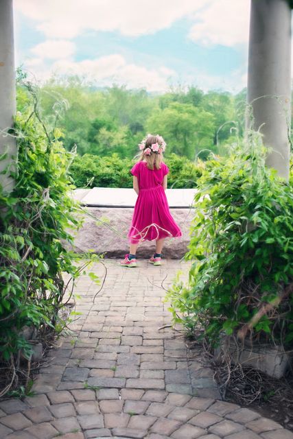 A young girl wearing a pink dress and flower crown is standing between two stone columns in a lush garden. She is facing away, looking at the greenery and landscape beyond. This image is ideal for use in content related to childhood, nature, carefree summer days, and outdoor activities.