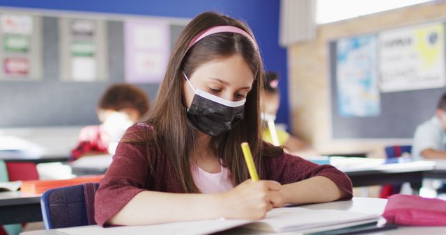 Young girl wearing a mask writing notes in a classroom. Ideal for topics on children's education, health and safety in schools, remote learning adaptations, pandemic impact on students, or academic-focused activities for children.