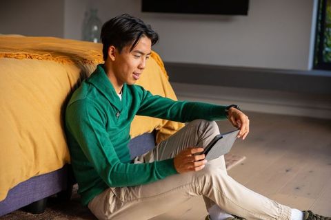 Asian man using tablet while sitting on bedroom floor wearing green sweater, cozy sunlit scene