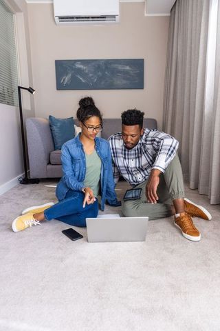 Young couple collaborating on laptop and tablet on carpeted living room floor