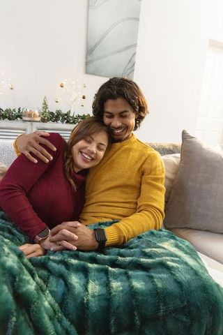 Cozy indian couple snuggling on sofa under blanket wearing mustard and burgundy sweaters