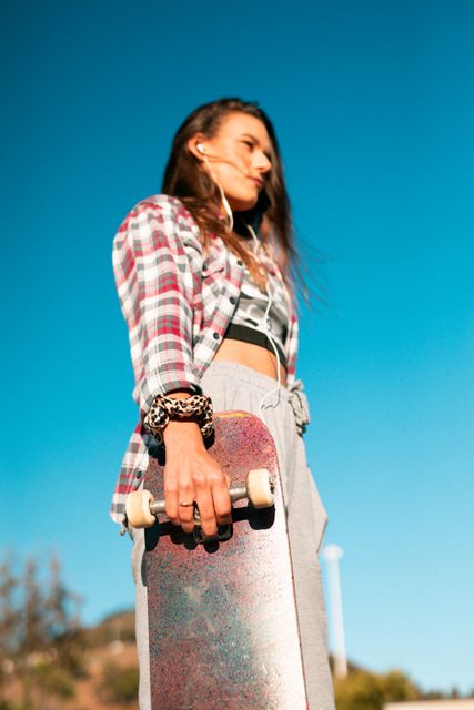 Caucasian woman wearing earphones, standing, holding skateboard in the sun - Download Free Stock Photos Pikwizard.com