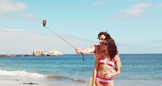 Couple wearing swimsuits taking a selfie with a selfie stick while standing on a beach. They are standing close to the ocean, with bright blue sky and ocean in the background. Ideal for use in travel advertisements, summer vacation promotions, or social media content about beach trips and leisure activities.