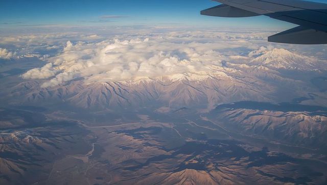 Flying over snow-dusted mountain range with airplane wing, clouds and winding riverbeds