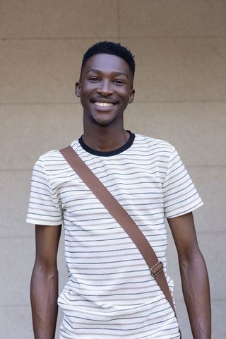 Smiling young man standing with crossbody strap against beige wall casual lifestyle portrait