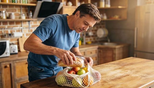 Man packing baguette into mesh bag while filling produce on rustic wooden kitchen island