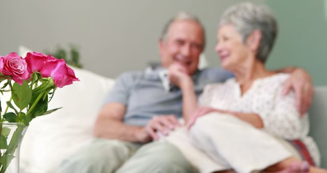 Elderly couple enjoying time together on sofa with pink roses in foreground. Perfect for articles about senior living, romance in old age, healthy aging, home decor, and lifestyle blogs centered around older adults. The image evokes feelings of comfort, togetherness, and sentimental love.