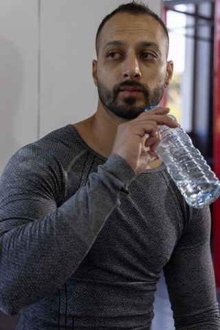 Mid-adult indian man sipping water after gym workout in fitted grey compression top, calm