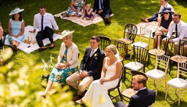 Sunlit garden wedding ceremony bride and groom sitting on wooden chairs and picnic guests