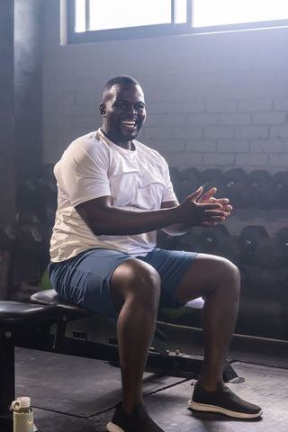 African american man clapping on bench in gym with dumbbells and backlit window