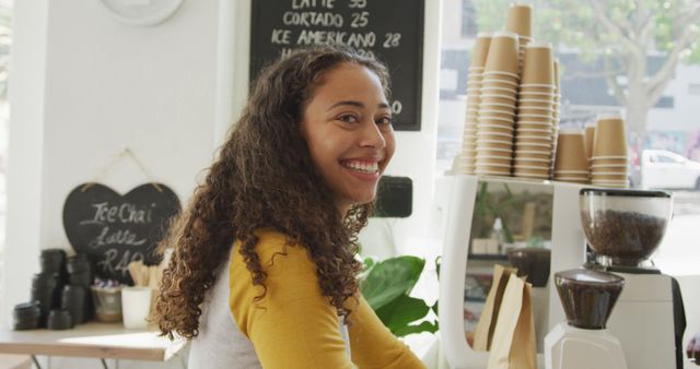 Young female barista is smiling while working behind the counter in a cozy, modern coffee shop. Ideal for content related to customer service, coffee shops, or young professionals in the food and beverage industry.