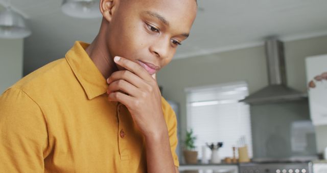 Young man standing in a modern kitchen, looking reflective and contemplative. He is casually dressed in a mustard-colored shirt. Ideal for concepts related to thinking, problem-solving, and moments of deep reflection. Suitable for use in blogs, advertisements, and articles focusing on personal development, mental health, and lifestyle topics.