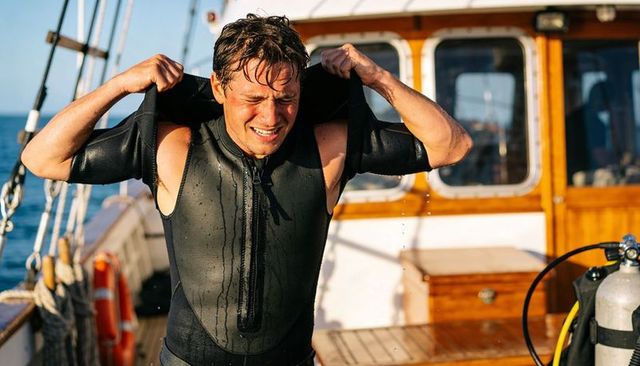Male diver pulling wetsuit top off on sunlit wooden dive boat deck with scuba tank