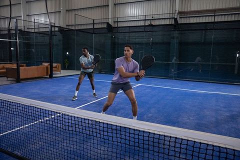 Two male teammates practicing doubles padel on indoor blue court holding rackets