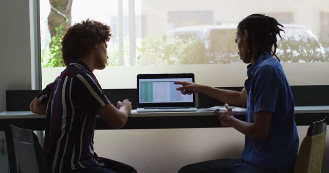 This image shows young colleagues engaged in reviewing data on a laptop in a modern office environment. It portrays teamwork and collaborative efforts in a casual yet professional setting. This can be useful for illustrating themes of workplace collaboration, tech workplace environments, data analysis, and modern office culture.
