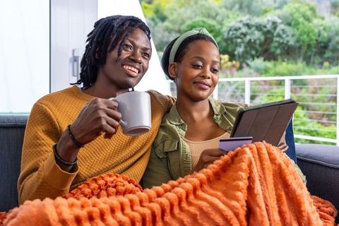 African american couple relaxing on sofa with tablet and credit card, cozy home shopping