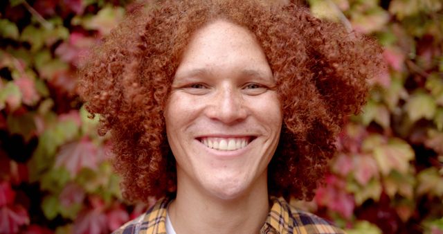 Young man with curly hair smiling warmly against background of autumn ivy leaves. His joyful expression and natural environment create positive and vibrant mood. Ideal for use in promotional materials, ads focused on youth and happiness, nature and outdoor themes, or mental well-being campaigns.