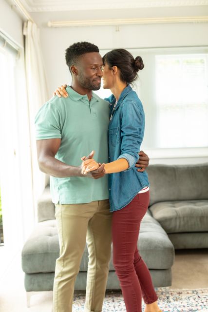 Young multiracial couple enjoying a romantic moment dancing together in their living room. Perfect for themes related to love, relationships, home life, and leisure activities. Ideal for use in lifestyle blogs, relationship advice articles, and home decor advertisements.