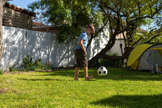 Caucasian boy kicking a ball around in their back yard. in the background is a tree with a tent set up beside it.