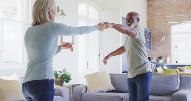 This image showcases a senior couple dancing happily in their modern, well-lit living room. They appear joyful and engaged in the moment, highlighting themes of love, togetherness, and an active lifestyle. Ideal for use in retirement planning, active aging content, home care services, and senior living advertisements and articles. The casual, natural setting with the couple's joyful expressions can appeal to audiences looking to highlight positive and affectionate aspects of senior life.