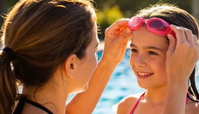Mother fitting pink goggles on smiling daughter at sunny pool building swim confidence