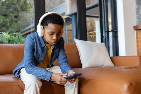 Preteen african american boy sitting on couch using headphones and smartphone concentrating