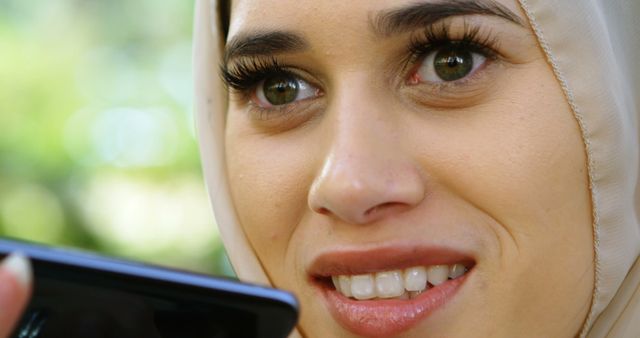 A close-up shot of a young woman wearing a hijab, smiling while using a smartphone. Best used for topics on modern technology, communication, lifestyle, and multiculturalism. Ideal for articles, advertisements, and educational materials highlighting technology's impact on everyday life.