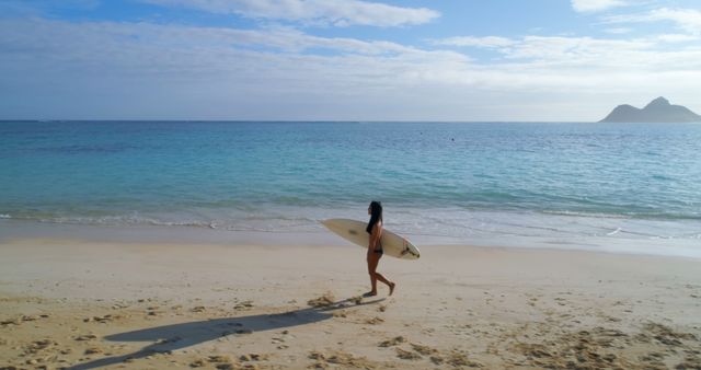 Young biracial woman walks on the beach holding a surfboard. She's ready for a surfing session in this serene outdoor setting.