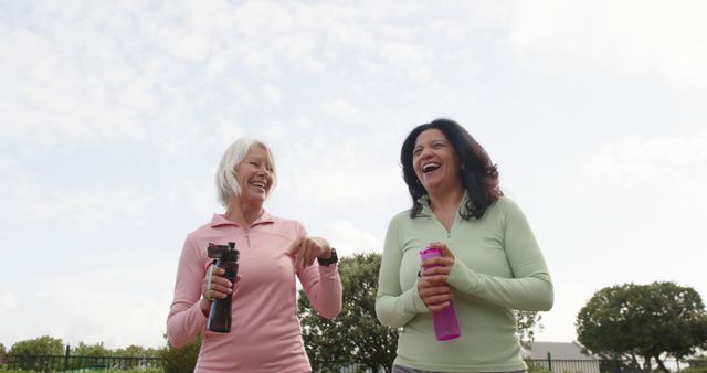 Two mature women are seen laughing while engaging in outdoor exercise. One woman holds a water bottle and the other a fitness gadget, reflecting an active and healthy lifestyle. Ideal for portraying themes of friendship, wellbeing, fitness in later life, and outdoor activities.