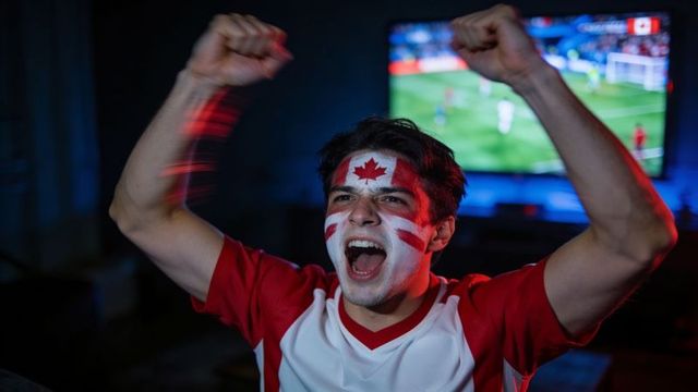 Canadian soccer fan cheering with maple leaf face paint watching match on tv at home