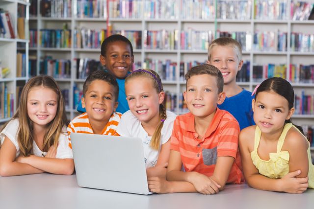 Diverse group of school kids gathered around a laptop in a library, smiling and engaged in learning. Ideal for educational content, school promotions, technology in education, and multicultural teamwork themes.