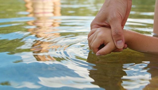 Adult Holding Child Hand Touching Calm Lake Water Ripples Reflecting Sky Bonding Moment