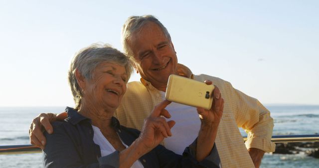 A senior couple are taking a selfie by the beach, smiling and enjoying their time together. This can be used for depicting themes of love, happiness, technology usage by seniors, retirement life, travel, and leisure.