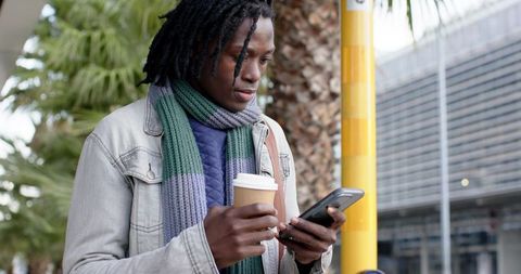 African american man checking phone while holding coffee at bus stop, urban commuter scene