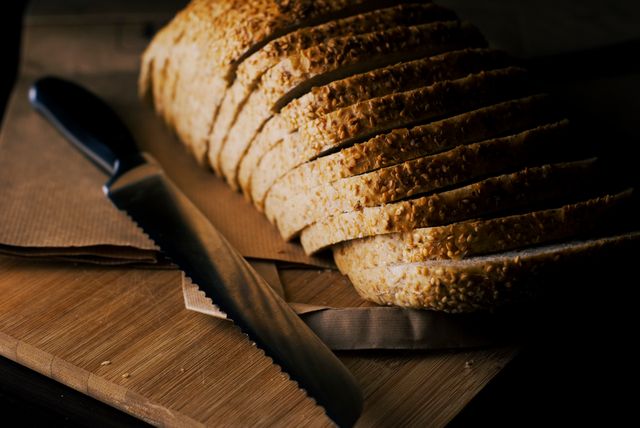 Freshly sliced loaf of bread with sesame seeds placed on a wooden cutting board. A bread knife sits beside it. Ideal for use in food blogs, recipe websites, bakery advertisements, or home cooking magazines featuring articles on homemade bakery products and breakfast ideas.