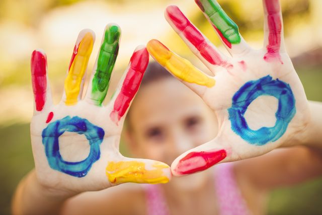 Child displaying brightly painted hands in a park, emphasizing creativity and playful expression. Ideal for use in educational materials, children's activities, art projects, and advertisements promoting creativity and outdoor play.