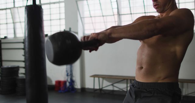 A muscular man engaging in strength training using a kettlebell inside a gym. He is shirtless, emphasizing his physical fitness and dedication to a healthy lifestyle. The image can be used in fitness blogs, training manuals, gym promotional materials, and health-related articles to illustrate physical fitness and motivation.