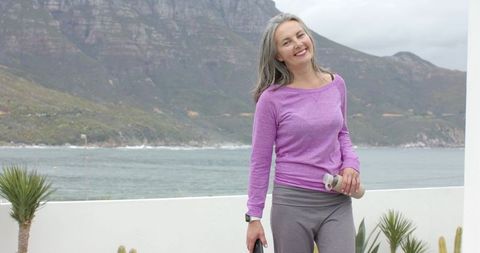 Smiling mature woman standing on coastal balcony holding reusable bottle embracing wellness
