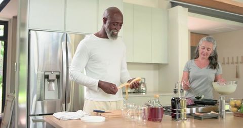 Mature diverse couple cooking breakfast on modern kitchen island peeling banana and whisking batter