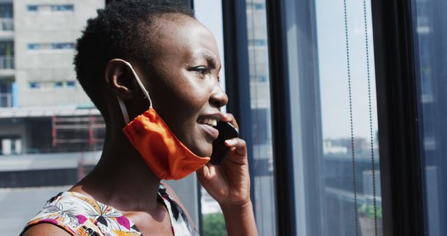 African woman smiling and conversing on phone near office window, suggesting a professional setting. Useful for topics related to business, communication, technology, and professionalism.