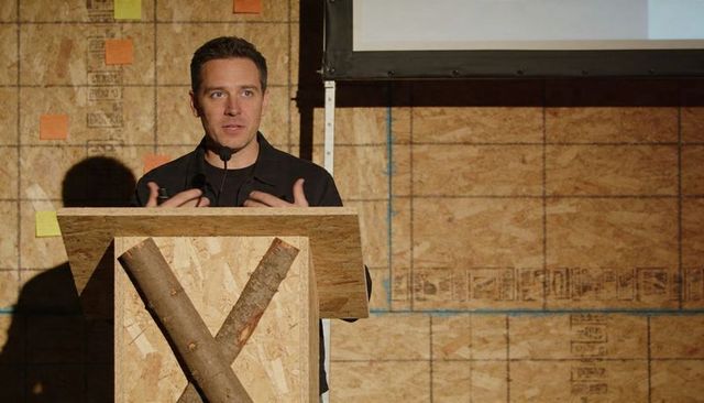 Male presenter gesturing at rustic wooden lectern with log x and projection screen onstage