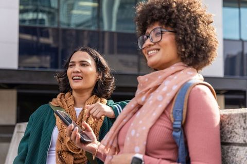 Two young women laughing and checking phone in urban plaza, wearing scarves and backpacks