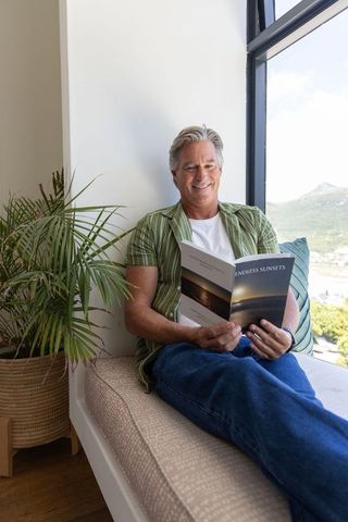 Senior man relaxing on sunlit window seat reading paperback about coastal sunsets