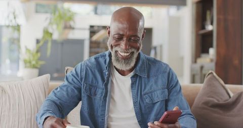 Senior african american man smiling while holding smartphone and mug on cozy sofa