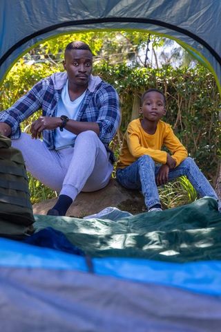 African american father and son sitting at tent entrance camping in sunlit woodland